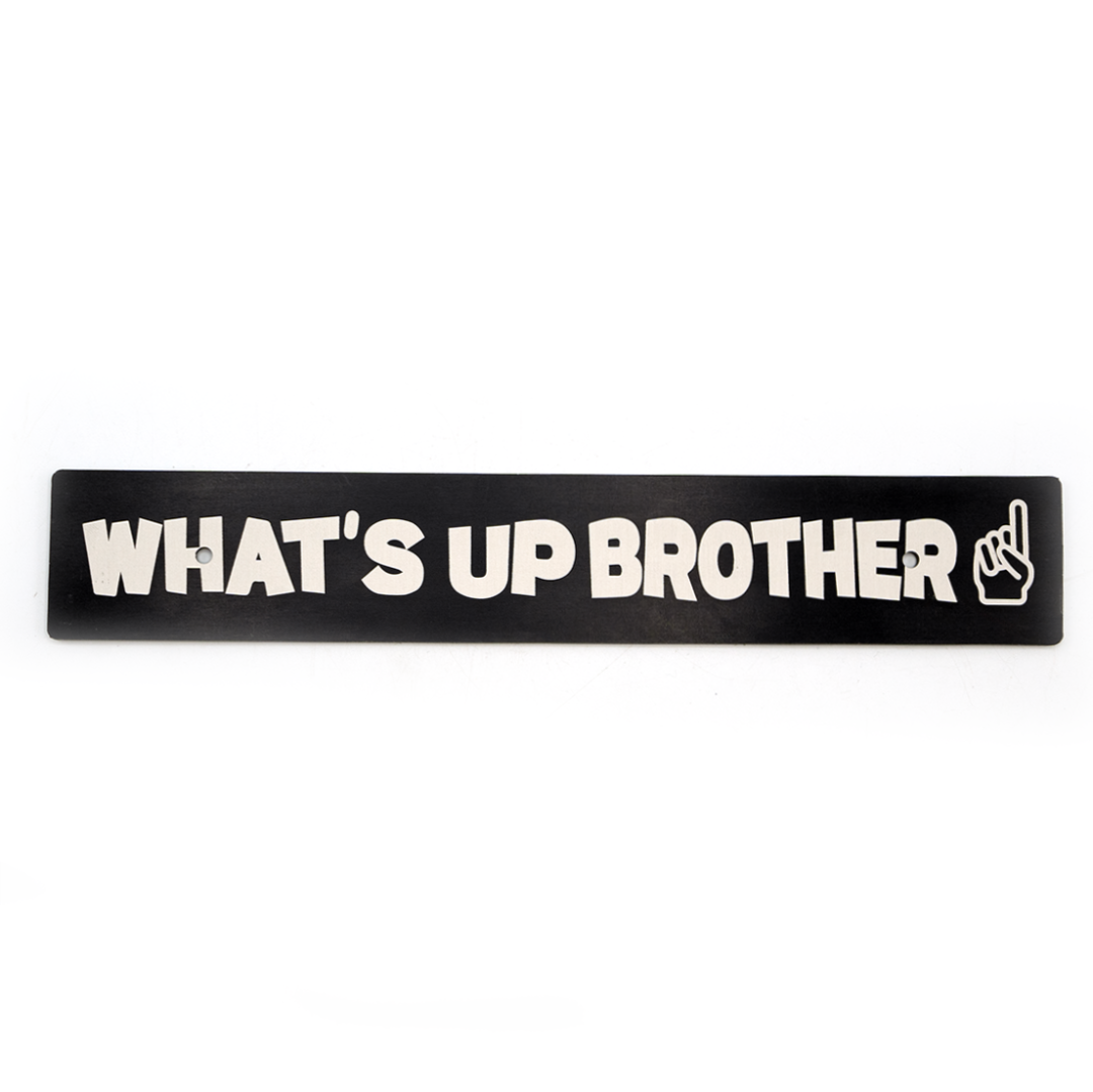 A black anodized aluminum license plate delete with white text 'WHAT'S UP BROTHER' and a pointing finger icon.