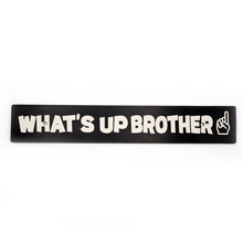 A black anodized aluminum license plate delete with white text 'WHAT'S UP BROTHER' and a pointing finger icon.