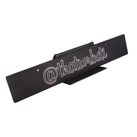Custom license plate delete with Instagram handle engraved in white, mounted on a black surface.