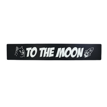 A black anodized aluminum front license plate delete with 'TO THE MOON' text and a rocket illustration in white.